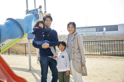 公園の滑り台の隣りで、娘さんを抱きかかえた旦那さん、息子さん、奥さんが並んで立っている家族写真