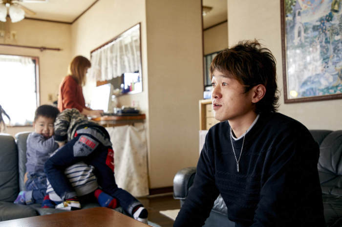 ソファに腰掛けている横田さん（右側）、ソファで団子状になって遊ぶ子ども3人（左側）、立って作業をしている奥さん（左奥）の写真
