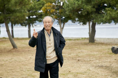 水辺の林で、右手人さし指を立て、左手をポケットに入れ、ポーズをとっている青山さんの写真